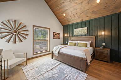 Cozy rustic bedroom with vaulted wood ceiling and dark wood-paneled accent wall, wooden king bed with green pillows, hanging pendant lights, nightstand, patterned area rug, white accent chair, metal windmill wall decor and window with blinds.
