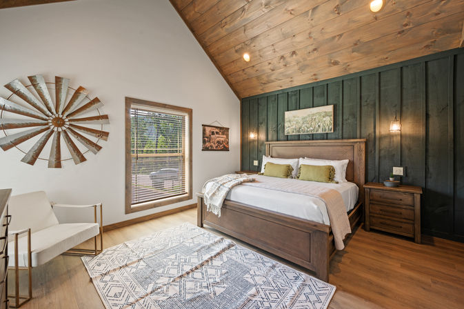 Cozy rustic bedroom with vaulted wood ceiling and dark wood-paneled accent wall, wooden king bed with green pillows, hanging pendant lights, nightstand, patterned area rug, white accent chair, metal windmill wall decor and window with blinds.