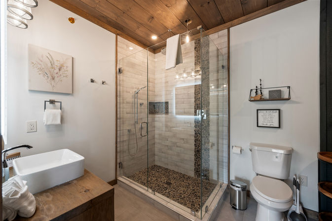 Modern rustic home bathroom with glass-enclosed walk-in shower, pebble stone shower floor and vertical stone accent, white vessel sink on a wooden vanity, wood‑paneled ceiling, towel rack and toilet — spa-like neutral design.