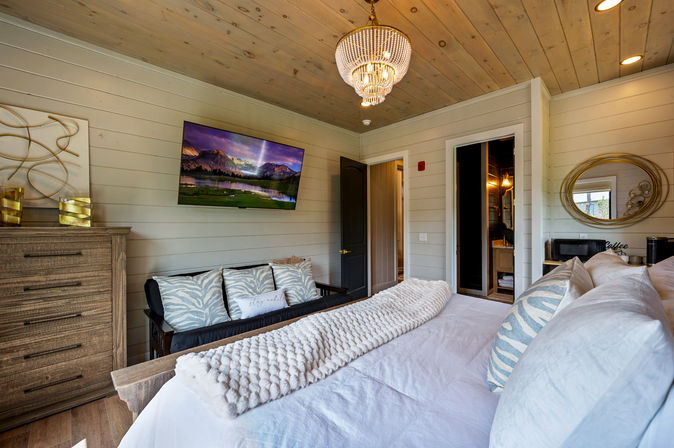 Cozy rustic bedroom interior with wood-plank ceiling and shiplap walls, crystal chandelier, wall-mounted TV, wooden dresser, sofa with patterned pillows, and a white bed topped with a plush knit throw.