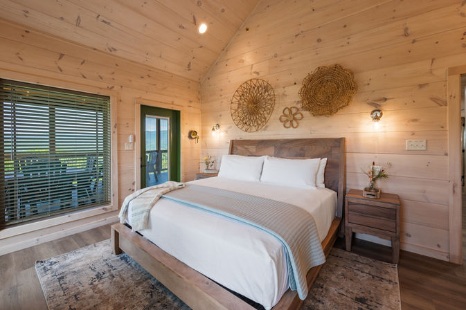 Bright, cozy rustic cabin bedroom — king bed with white linens, wood-paneled walls, woven wall art, bedside tables and a glass door opening to a porch with a scenic outdoor view.