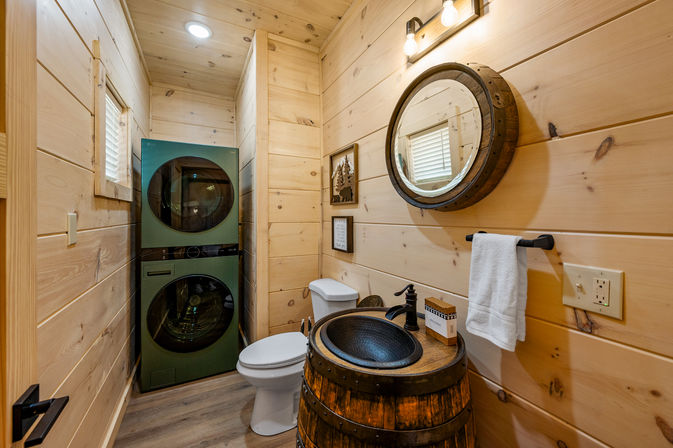 Cozy cabin-style bathroom with stacked green washer/dryer, white toilet, barrel vanity with black vessel sink, round wooden mirror and pine plank walls.