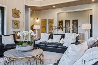 Bright modern living room with dark leather sofa, neutral pillows and knit throws, round stone-top coffee table with white orchids, brass-accent base, wood-paneled walls, gold wall art and warm table lamps.