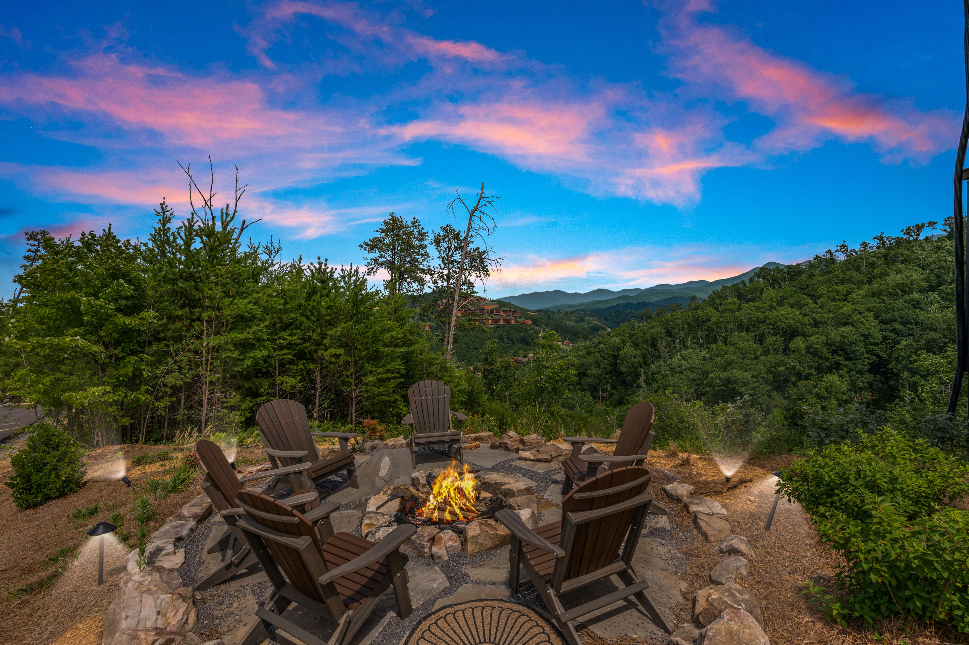 Cozy stone fire pit surrounded by six Adirondack chairs on a forested mountain overlook under a pink-and-blue sunset sky