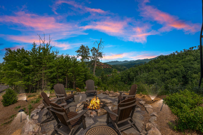 Cozy stone fire pit surrounded by six Adirondack chairs on a forested mountain overlook under a pink-and-blue sunset sky