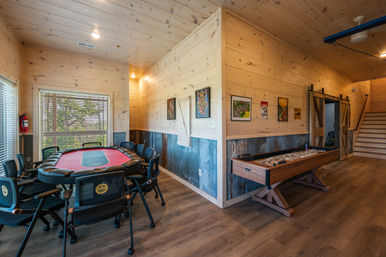 Rustic cabin game room with red-felt poker table surrounded by chairs, wooden shuffleboard table, light wood-paneled walls with corrugated metal wainscoting, barn-style sliding door, and large window with natural light.