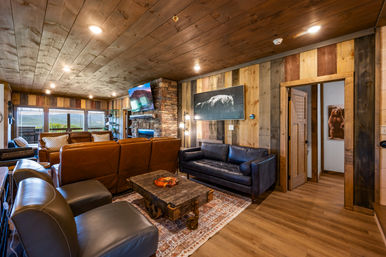 Cozy rustic mountain-cabin living room with stone fireplace, leather sofas and armchairs, reclaimed wood walls and ceiling, a vintage wooden coffee table, and large windows with valley views.