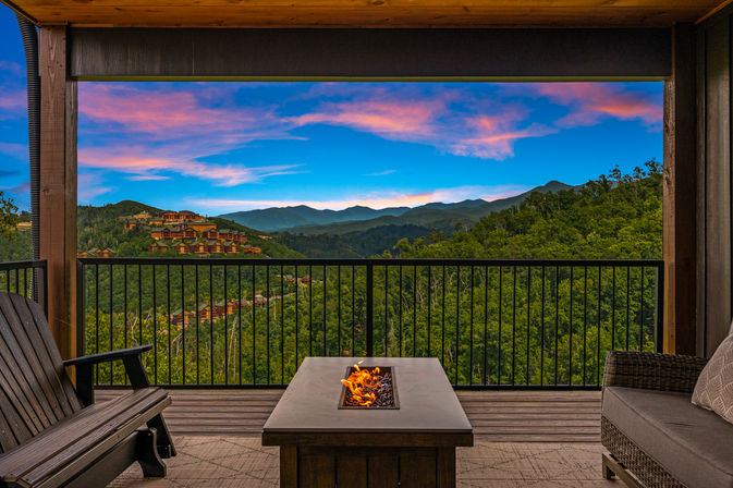 Covered wooden deck balcony with seating and a fire-pit table, framing a panoramic view of lush forested mountains, hillside cabins, and a vivid pink-and-blue sunset sky.