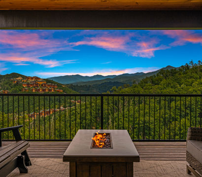 Covered wooden deck balcony with seating and a fire-pit table, framing a panoramic view of lush forested mountains, hillside cabins, and a vivid pink-and-blue sunset sky.
