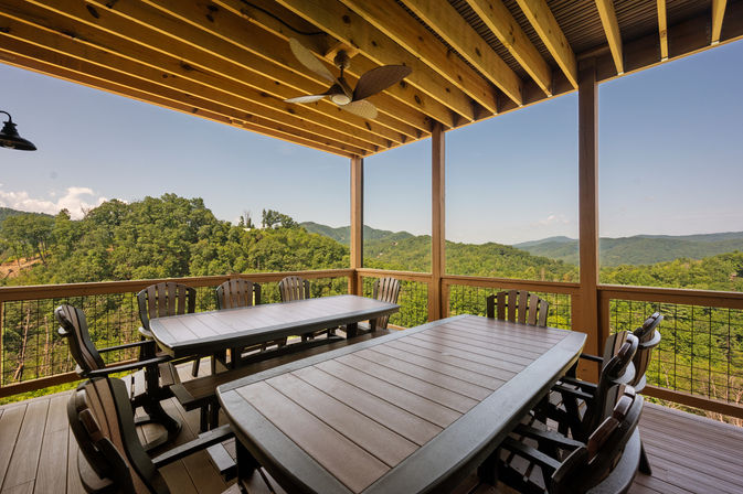 Scenic covered wooden deck with ceiling fan and two long outdoor dining tables and chairs, overlooking lush forested hills and distant mountain range under a clear blue sky