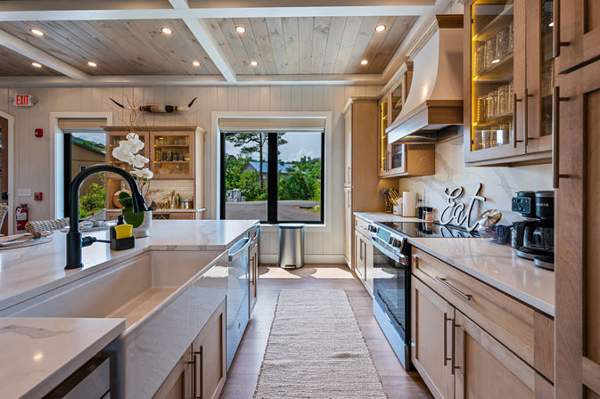 Bright modern farmhouse kitchen with a large island and farmhouse sink, marble countertops, light wood cabinets, stainless appliances, recessed ceiling lights, and a central window overlooking greenery.
