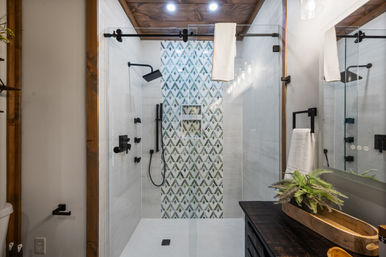 Modern farmhouse bathroom shower with sliding glass barn-door, geometric green-and-white accent tile, matte black fixtures, wood ceiling trim and dark vanity with potted fern.