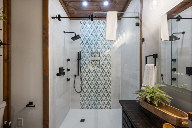 Modern farmhouse bathroom shower with sliding glass barn-door, geometric green-and-white accent tile, matte black fixtures, wood ceiling trim and dark vanity with potted fern.