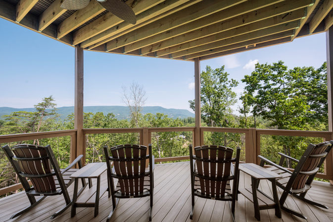 Covered wooden deck with ceiling fan, four Adirondack-style chairs and side tables facing a green forest and distant blue mountains under a clear sky