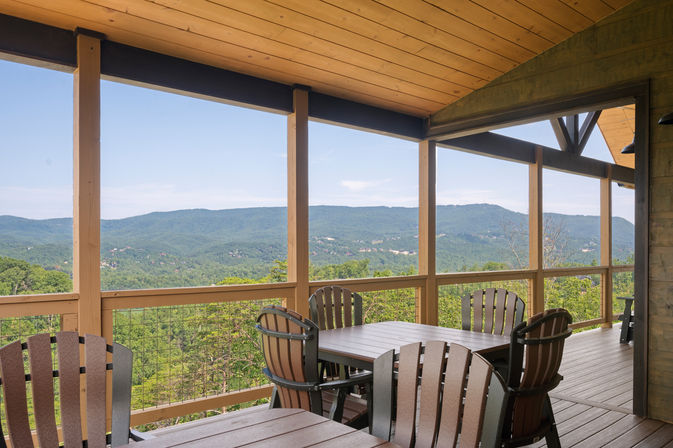 Covered wooden deck with outdoor dining table and Adirondack-style chairs overlooking a panoramic green mountain view and forested valley.