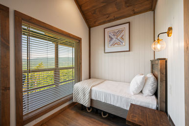 Cozy rustic single bedroom in a mountain cabin with white paneled walls, wood trim and ceiling, a neatly made twin bed with quilt and pillows, brass globe wall sconce, and a large window with blinds framing a green mountain view.