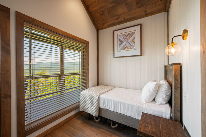 Cozy rustic single bedroom in a mountain cabin with white paneled walls, wood trim and ceiling, a neatly made twin bed with quilt and pillows, brass globe wall sconce, and a large window with blinds framing a green mountain view.