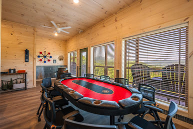 Cozy wood-paneled mountain cabin game room with a red-felt poker table surrounded by black chairs, large windows with blinds revealing forested hills and mountain views, outdoor deck seating, ceiling fan and rustic decor.