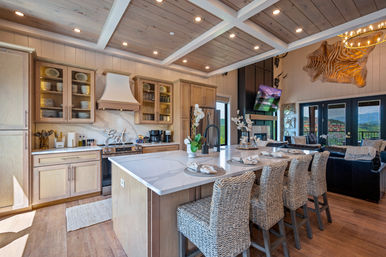 Bright open-concept mountain home kitchen with marble-topped island, four woven rattan bar stools, light wood cabinetry, coffered wood ceiling, stainless range with decorative "Eat" sign, and adjoining living room with fireplace, wall TV and large glass doors revealing panoramic mountain views.