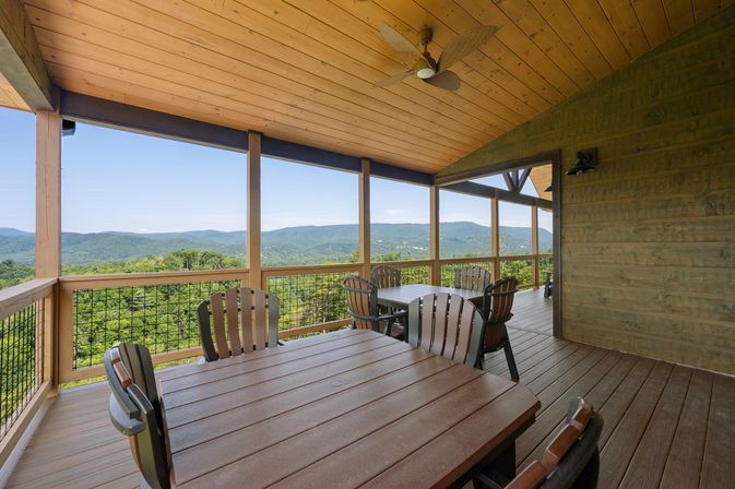Covered wooden deck with ceiling fan, patio dining tables and chairs overlooking forested hills and distant mountains — scenic mountain view porch.