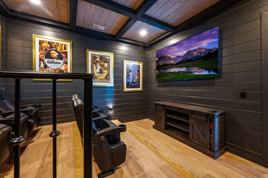 Cozy home theater room with dark shiplap walls and wood‑beamed ceiling, wall‑mounted TV displaying a mountain lake, leather recliner seats, framed movie posters, and a rustic wooden media console on hardwood floors.