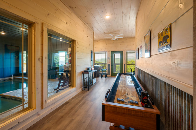 Cozy cabin-style game room with a shuffleboard table, pine plank walls and ceiling, large interior windows revealing an indoor pool, and a glass door with a scenic outdoor view.