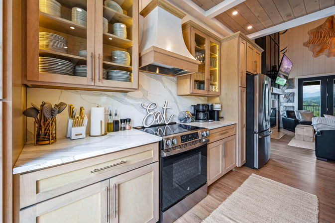 Sunlit modern farmhouse kitchen with light wood cabinets, marble backsplash and countertops, stainless steel range and fridge, glass-front dish cabinets, ‘Eat’ sign on the stovetop, coffee station and open-concept living area with mountain views.