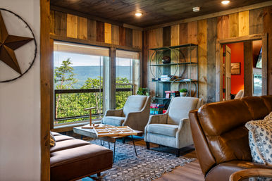 Cozy rustic mountain cabin living room with wood-paneled walls, two gray armchairs and leather sofa around a game-topped coffee table, circular shelf with decor, patterned rug, and a large window framing tree-covered hills and distant mountains.