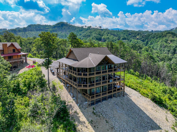 Multi-level mountain lodge with wraparound balconies perched on a forested hillside under bright blue skies, offering panoramic mountain views