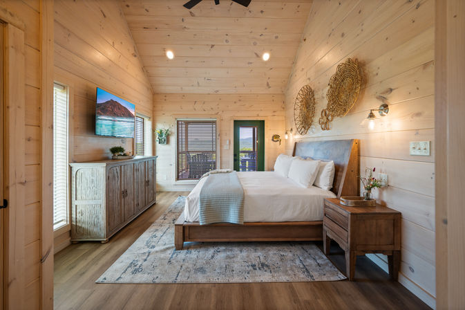 Cozy sunlit rustic wooden cabin bedroom with king bed, woven wall decor, wooden nightstand and dresser, wall-mounted TV, area rug, and glass door opening to a balcony with distant mountain view.