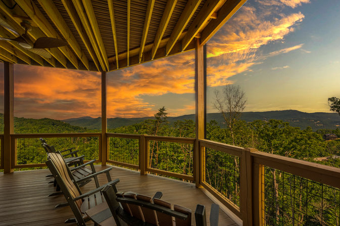 Cozy covered wooden deck with Adirondack chairs overlooking forested rolling mountains at sunset, dramatic orange clouds stretching across the sky.