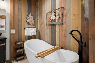 Cozy rustic bathroom with a white freestanding soaking tub, wooden bath caddy, black floor-mounted faucet, reclaimed wood plank accent wall, towel rack and metal tree wall art.
