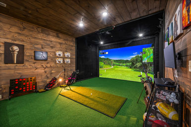 Cozy wood-paneled home indoor golf simulator with large projector screen showing a green fairway, artificial turf hitting mat, golf bags and clubs, wall art and sports gear