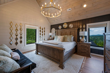Cozy rustic-chic master bedroom with vaulted shiplap ceiling, wooden king bed and patterned pillows, candle-style chandelier, gold decorative mirrors, bedside lamps, textured rug and twin windows framing lush green mountain views.