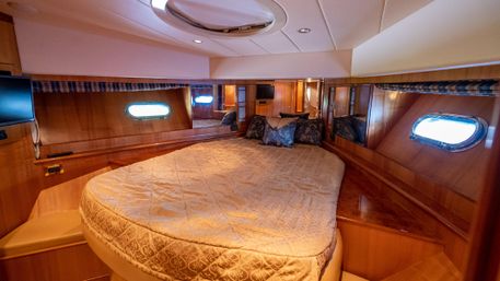 Wood-paneled yacht cabin interior with rounded queen bed, beige patterned comforter, blue decorative pillows, oval portholes, mirrored walls and ceiling hatch