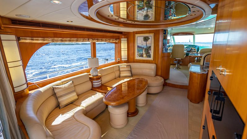 Sunlit luxury yacht salon with curved beige leather sofa, glossy oval wood table, mirrored ceiling detail, framed artwork and panoramic ocean view through large windows toward the helm