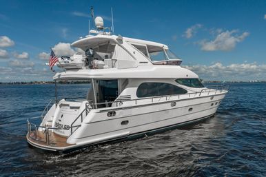Sleek white luxury motor yacht gliding on calm coastal waters under a bright blue sky, American flag flying at the stern, flybridge with dinghy and radar, teak swim platform visible.
