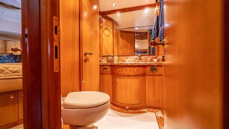 Warm wood-paneled luxury yacht bathroom with white porcelain toilet, marble vanity and mirror, blue hand towels and adjoining cabin visible
