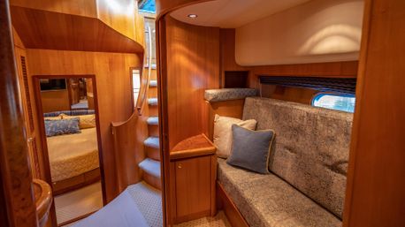 Cozy wood-paneled luxury yacht cabin interior with curved staircase, built-in patterned upholstered sofa and throw pillows, small porthole window and a bedroom visible through the doorway.