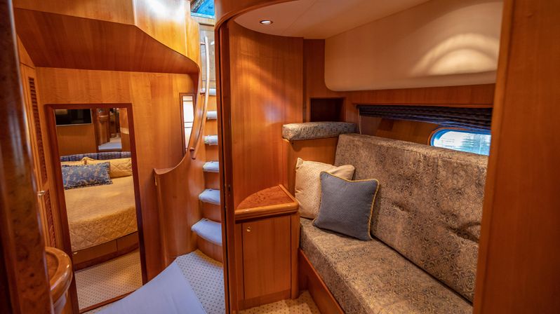 Cozy wood-paneled luxury yacht cabin interior with curved staircase, built-in patterned upholstered sofa and throw pillows, small porthole window and a bedroom visible through the doorway.