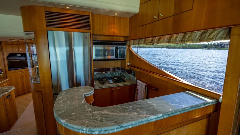 Luxury yacht galley with curved green marble countertop, stainless-steel refrigerator, built-in microwave and stovetop, and a large panoramic side window overlooking calm coastal water.