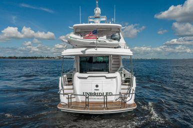 Rear view of a white luxury motor yacht with inflatable tender and American flag, cruising on blue ocean waters under a sunny sky with puffy clouds near a low coastal shoreline
