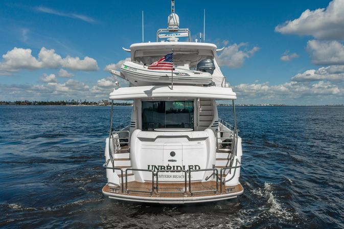 Rear view of a white luxury motor yacht with inflatable tender and American flag, cruising on blue ocean waters under a sunny sky with puffy clouds near a low coastal shoreline