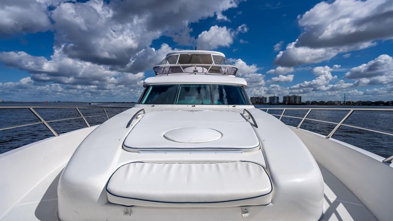 Front view of a white luxury yacht bow with cushioned sun pad and stainless railings on calm coastal waters under a bright blue sky with dramatic clouds and a distant shoreline skyline.