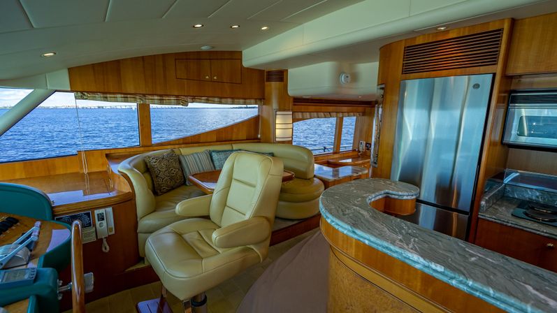 Sunlit luxury yacht interior: cream leather captain’s chair and curved dinette with patterned pillows, marble-topped galley counter, stainless-steel refrigerator, warm wood paneling and large windows overlooking the ocean.