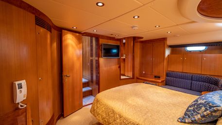 Cozy luxury yacht cabin — queen bed with quilted gold cover, warm wood paneling, navy patterned settee, wall-mounted TV, porthole window and small steps to the companionway.