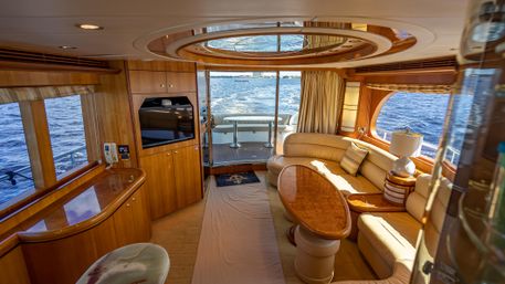 Sunlit luxury yacht salon at sea with curved beige leather seating, glossy wood coffee table and cabinetry, panoramic windows, skylight and aft deck view of the open ocean