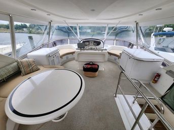 Enclosed yacht flybridge with central helm and navigation screens, wraparound windows overlooking a marina, curved cushioned seating, white oval table, storage cabinets and stainless railings.