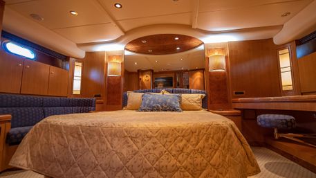 Luxury yacht master stateroom with queen bed and decorative pillows, wood-paneled walls, blue upholstered seating, porthole window and warm ambient lighting