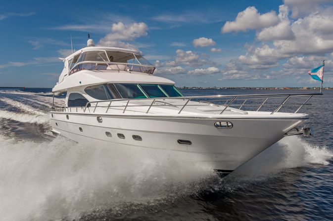 White luxury motor yacht cruising on blue coastal waters, bow cutting through waves with spray under a sunny sky dotted with puffy clouds.
